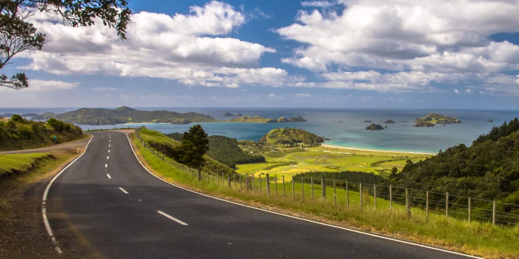 Inviting road through New Zealand countryside with blue sea with tropical islands at Bay of Islands, Northland, New Zealand