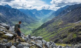 A man in his mid-20's hikes with a daypack down a steep trail overlooking the Little Susitna River in the Gold-Mint Trail Valley of the Hatcher Pass area in the Talkeetna Mountain Range of Alaska.