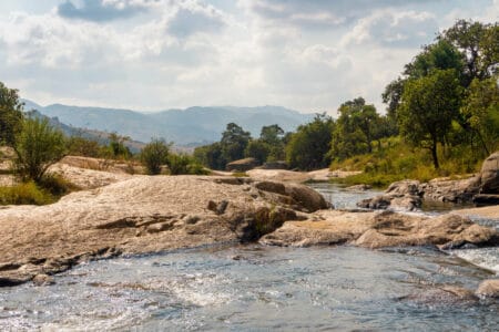 Felsiger Flusslauf mit klarem Wasser, grüner Vegetation und Bergen im Hintergrund unter leicht bewölktem Himmel in Eswatini, Südafrika.